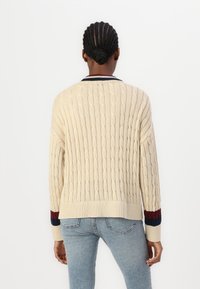 Pull crème en maille torsadée avec un ourlet côtelé et des accents rayés bleu marine et rouge sur les manches. Présente une coupe décontractée et un motif texturé.