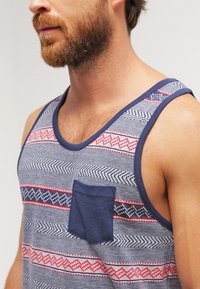 Débardeur rayé bleu marine et gris en coton, avec une poche poitrine et des motifs géométriques rouges et bleus sur le tissu.