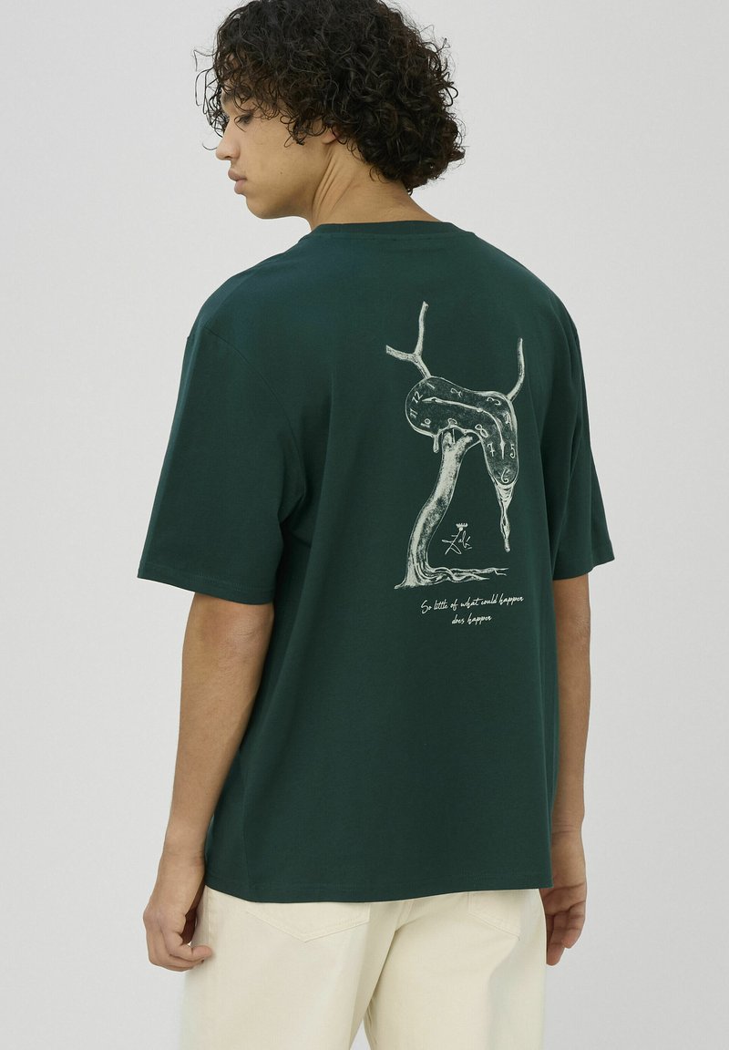 Jeune personne aux cheveux bouclés portant un t-shirt vert foncé avec un motif d'horloge fondante et un pantalon de couleur claire, vue de dos.