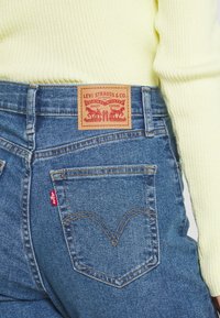 Primer plano del bolsillo trasero de unos jeans Levi's azules y la etiqueta, llevados con un suéter amarillo de canalé.