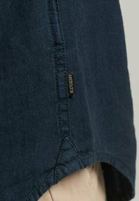 Camicia in lino blu navy con orlo curvo, caratterizzata da cuciture evidenti e un'etichetta del marchio discretamente posizionata di lato. Il tessuto presenta una finitura testurizzata.