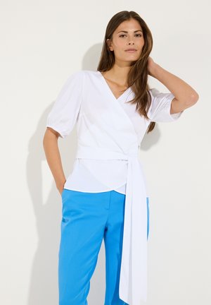 Frau mit langen braunen Haaren, die eine weiße Wickelbluse mit Puffärmeln und leuchtend blaue Hose trägt, posiert vor einem einfarbigen Hintergrund.