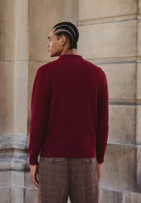 Pull en maille bordeaux avec une finition texturée, doté d'un col et de poignets côtelés, associé à un pantalon à carreaux marron.