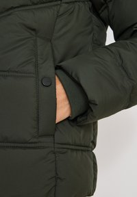 Mørkegrøn, polstret jakke med et nærbillede af en hånd i en sidelomme. Stoffet har en glat tekstur med en subtil glans.