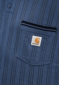 Μπλε πλεκτό ύφασμα με κάθετες διάτρητες ρίγες, με τσέπη με σκουρόχρωμη επένδυση και ετικέτα λογότυπου Carhartt.