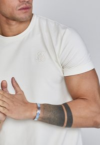 Camiseta blanca de manga corta con un logo texturizado "TS" en el pecho, acompañada de una pulsera plateada con acentos azules y un tatuaje en el brazo.