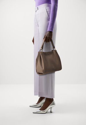 Braune Ledertasche mit weicher Textur, geometrischer Form und goldenen Hardware-Akzenten. Modell trägt helllilafarbene Hosen und silberne Absätze.