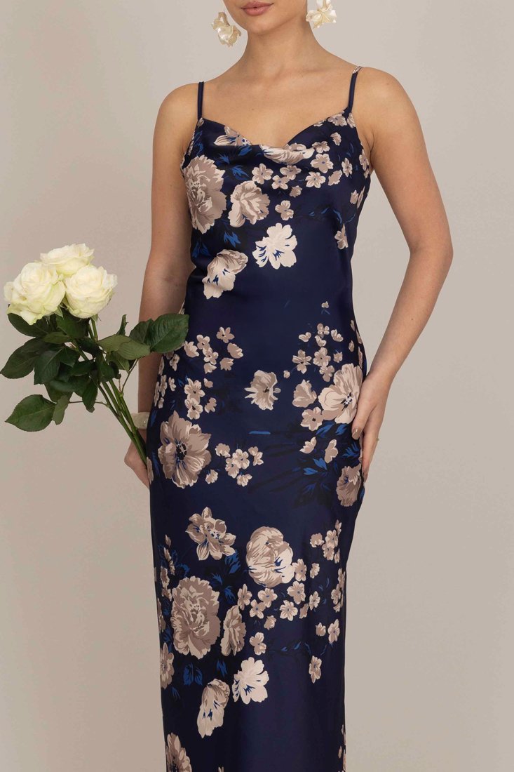 Abito in satin floreale di colore blu navy con fiori beige e blu. Vestibilità slim, scollatura a portafoglio e spalline regolabili. Il modello tiene in mano delle rose bianche.
