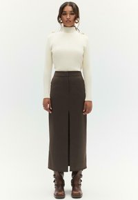 Witte gebreide trui met een hoge hals en ribbeltextuur, gecombineerd met een bruine, hoge taille rok met een split aan de voorkant, gedragen met bruine laarzen.