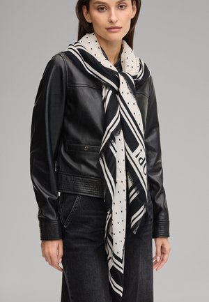 Frau trägt schwarze Lederjacke, schwarze Hose und einen großen weißen Schal mit schwarzen Punkten und Streifen, der um ihren Hals gebunden ist.