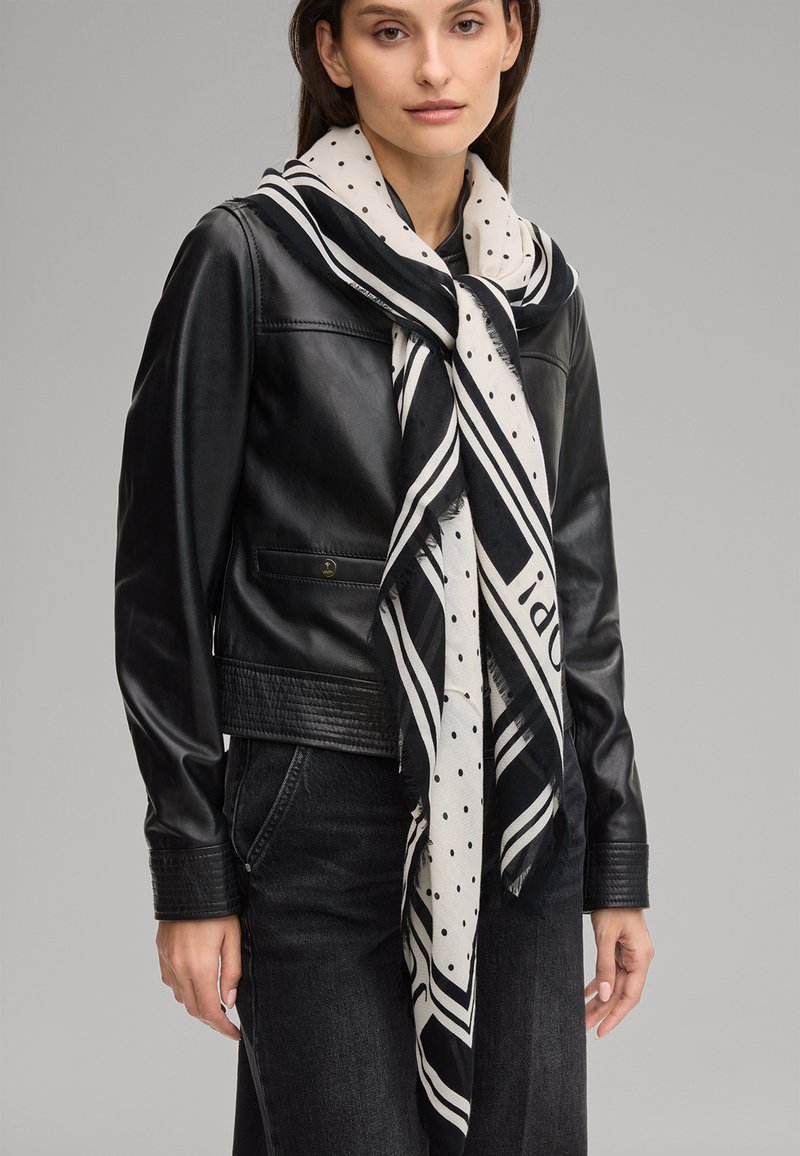 Frau trägt schwarze Lederjacke, schwarze Hose und einen großen weißen Schal mit schwarzen Punkten und Streifen, der um ihren Hals gebunden ist.