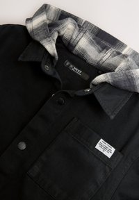 Camicia nera con bottoni e cappuccio in flanella a quadri grigia e bianca. Presenta una tasca frontale e un'etichetta sulla tasca.