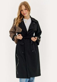 Čierny trenčkot s rukávmi s leopardím vzorom, opásaný v páse, dvojradový predok a bočné vrecká; obsahuje kontrastný biely text.