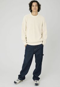 Pull enrique en côtelé crème à manches longues, associé à un jean en denim bleu foncé ample. Des baskets blanches et grises complètent la tenue.