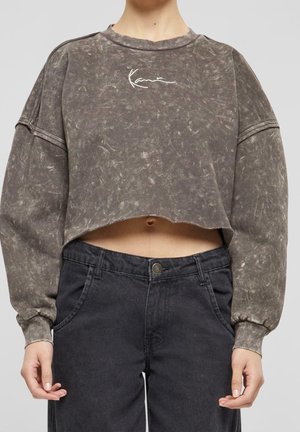 Persoon die een cropped, vergeeld grijs sweatshirt met lange mouwen en zwarte high-waist jeans draagt tegen een effen achtergrond.