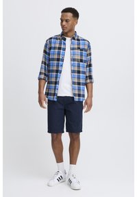 Plaid button-up shirt in blue, brown, and black, worn over a white T-shirt. Paired with navy shorts and white sneakers with black accents.