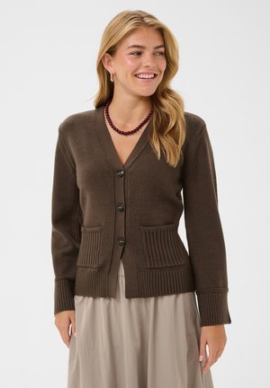Femme souriante, portant un cardigan marron boutonné avec deux poches avant et une jupe beige, accessoirisée d'un collier sombre à perles.