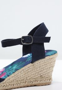 Sandale compensée en tissu bleu marine avec semelle intérieure fleurie, bride de cheville réglable, boucle métallique et talon en jute tressée.
