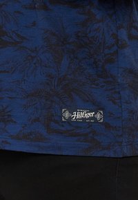 Plava tkanina s uzorkom crne palme, s bijelom oznakom koja u donjem kutu glasi "Hilfiger".