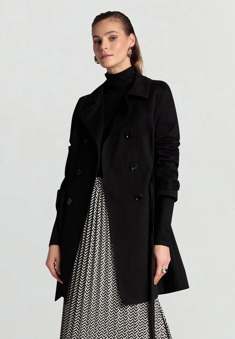 Frau trägt einen schwarzen doppelt geknöpften Mantel, schwarzen Rollkragenpullover, silberne Creolen, silbernen Ring und einen schwarz-weiß gemusterten Chevron-Rock.