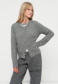 Grå strikket cardigan med rund hals, knappelukking, to frontlommer og en myk tekstur, brukt over grå cargo-bukser.