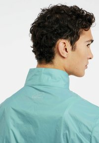 Šviesiai mėlynas švarkas, pagamintas iš sklandžios, lengvos medžiagos, su aukštu apykakle ir maža "PROTEST" logotipu ant nugaros.