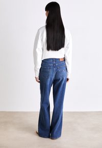 Pantalones vaqueros de denim azul desgastado, de pierna ancha y con una cintura de altura media, con cinco bolsillos y una etiqueta de cuero marrón en la parte posterior. Combinado con una blusa blanca.