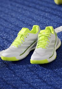 White athletic shoes with neon yellow accents and laces resting on blue textured sports court near white line.