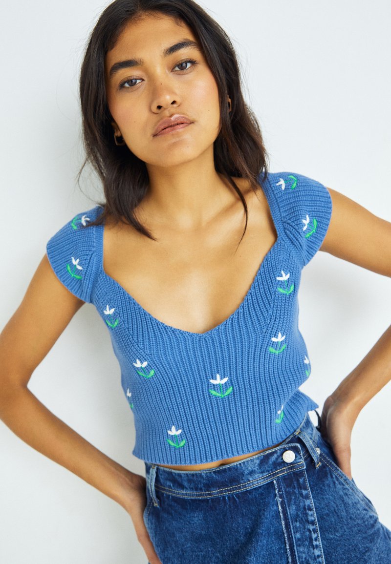 Kék kötött crop top mély V-nyakkivágással, rövid ujjakkal, fehér és zöld virágmintákkal. Farmer szoknyával viselve.