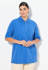 Modrá polo košile s límečkem, s krátkými rukávy a zapínáním na knoflíky. Tkanina vypadá hladce; model má na sobě neutrální kalhoty.