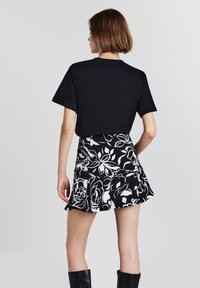 Femme aux cheveux bruns courts portant un t-shirt noir, une jupe florale noire et blanche, et des bottes noires jusqu'aux genoux, se tenant de dos.