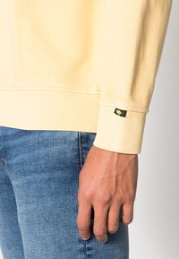 Sweat-shirt jaune clair avec une texture douce, doté d'une étiquette sur le poignet. Porté avec un jean en denim bleu, montrant le détail du bras et de la main.