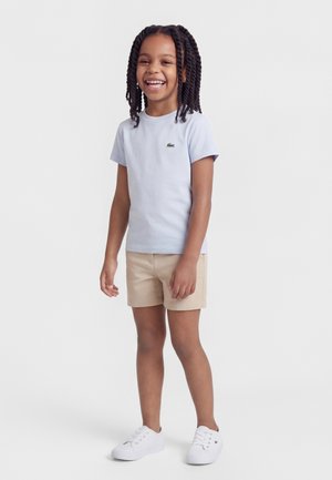 Smilende barn med flettet hår iført en lyseblå t-shirt, beige shorts og hvide sneakers, der står foran en ensfarvet baggrund.