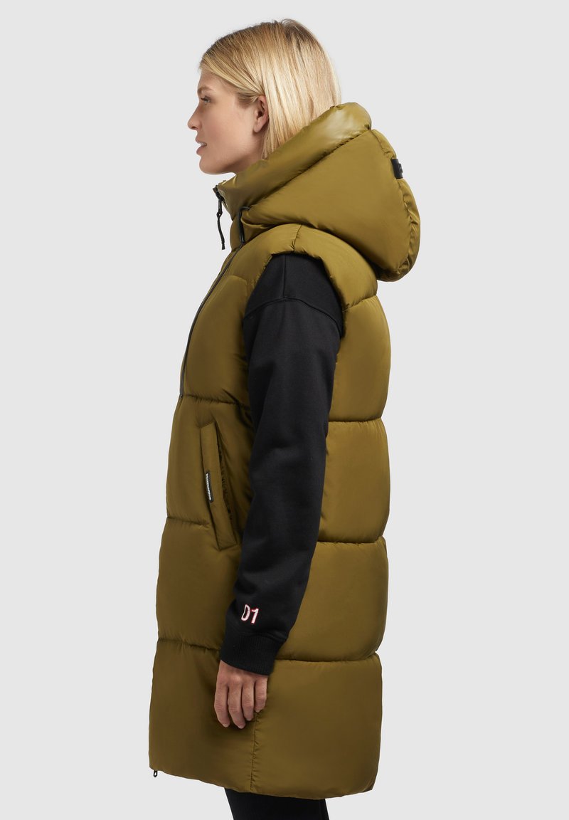 Frau mit blonden Haaren, die eine lange olivgrüne Steppweste mit Kapuze über einem schwarzen Langarmshirt trägt, steht im Profil vor einem grauen Hintergrund.