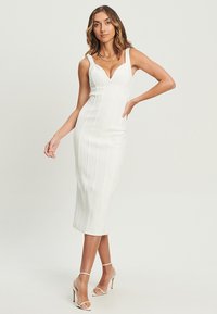 Witte aansluitende midi-jurk met smalle bandjes, gestructureerd lijfje en verticale naden, gecombineerd met transparante hoge hakken.