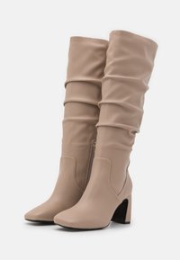 Beige knähöga stövlar med slouchig design, tillverkade av ett mjukt material. Har en sidodragkedja och en klack för stabilitet.