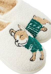 Pantofole bianche in peluche con motivo di un cane, con un pattern a maglione verde. Texture morbida e forma arrotondata con una fodera interna beige.