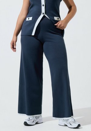 Gros plan d'une personne portant un pantalon large bleu marine, un gilet assorti avec bord blanc et des baskets blanches, debout sur un fond uni.