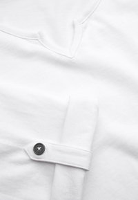 Hvid bomulds t-shirt med V-hals, glat tekstur og oprullede ærmer, som er sikret med en sort knap.