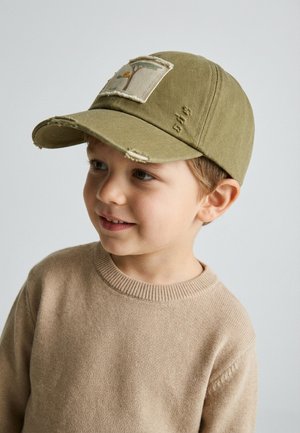 Junge lächelt, trägt einen beigen Pullover und eine olivgrüne Mütze mit einem Aufnäher von einem Vogel und Baum auf der Vorderseite.