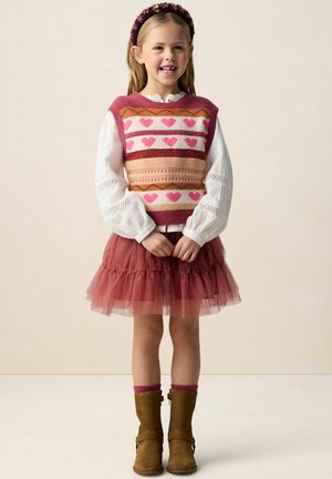 Jong meisje met een glimlach, gekleed in een roze spencer met hartjespatroon, een wit blouse met lange mouwen, een roze tule rok, bruine laarzen en een hoofdband met bloemen.