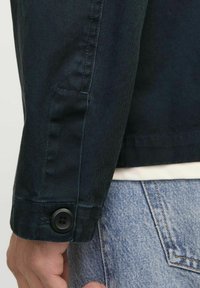 Zwarte spijkerjackmouw met een zwarte knoopsluiting. Kenmerkt zich door de textuurstof en een getailleerde fit. Gedragen met lichtblauwe jeans.
