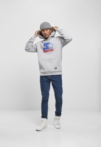 Grå hættetrøje med et blåt og rødt "STARTER" logo, parret med mørke jeans og hvide højtop sneakers, sat mod en ensfarvet baggrund.