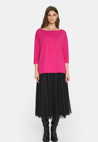 Fuksjowa bluzka z długim rękawem o szerokim dekolcie, zestawiona z czarną, warstwową spódnicą maxi z tiulu i czarnymi, wysokimi na kolana botkami.