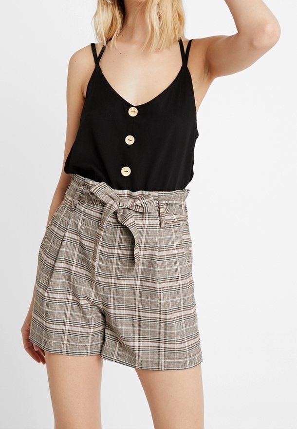 Top senza maniche nero con tre bottoni in legno; pantaloni corti a vita alta a quadri in beige, marrone e bianco, con cintura annodata e pieghe.