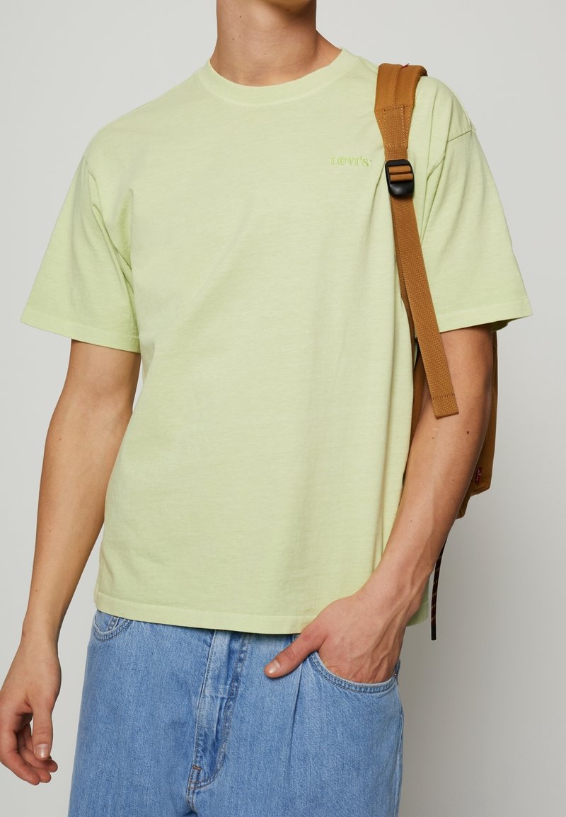 Ljusgrön kortärmad t-shirt med rund hals och Levi's-logotyp på vänster bröst, matchad med ljusblå jeans och en brun ryggsäckstropp.