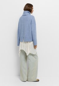 Világos kék, túlméretezett pulóver, magas bordázott gallérral, hátsó részen hasítékkal. Egy laza fehér struktúrájú felsőn és világos denim nadrágon viselve.