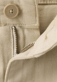 Gros plan sur un tissu beige montrant un bouton et une fermeture éclair argentée partiellement ouverte sur un matériau de vêtement plié.