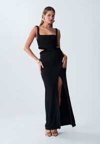 Black, form-fitting dress with a square neckline, ruching detail, side cutouts, and a thigh-high slit. Worn with gold accessories.