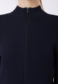 Marineblå ribbestrikket genser med høy krage og svart glidelås. Stoffet har en tekstur med vertikale linjer som skaper en figurnær silhuett.